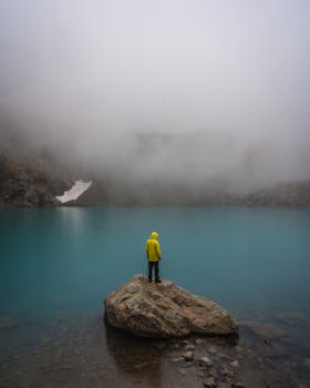 Person in a yellow jacket stands on a rock by a foggy blue lake, creating a serene and moody scene.