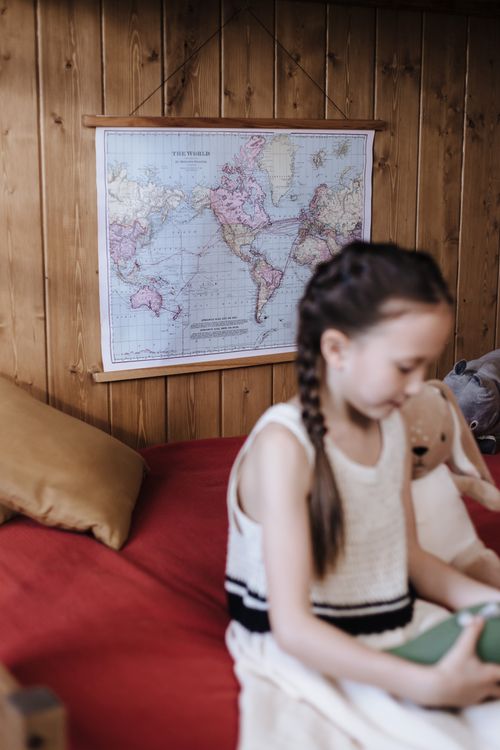 Free A young girl with braids reads indoors, seated on a bed with a world map in the background. Stock Photo