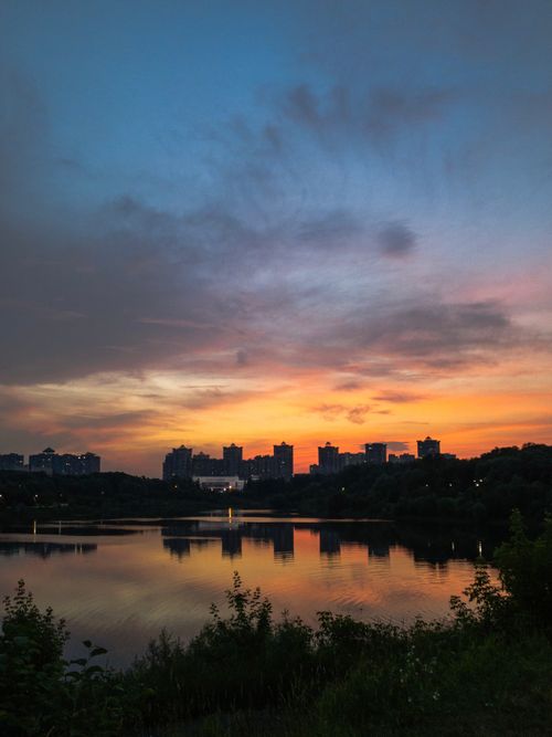 Free A tranquil sunset casts vibrant colors over a calm lake and silhouetted city buildings. Stock Photo