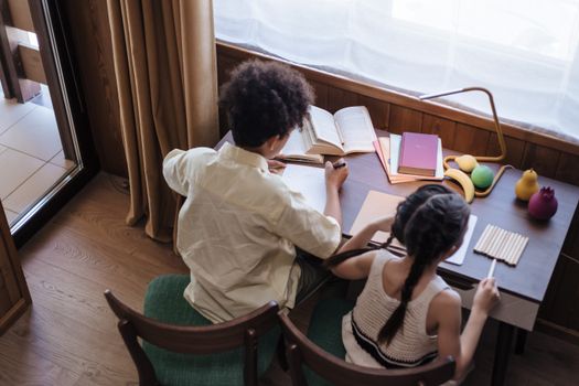 Two children studying with open books and stationery at home, viewed from above.