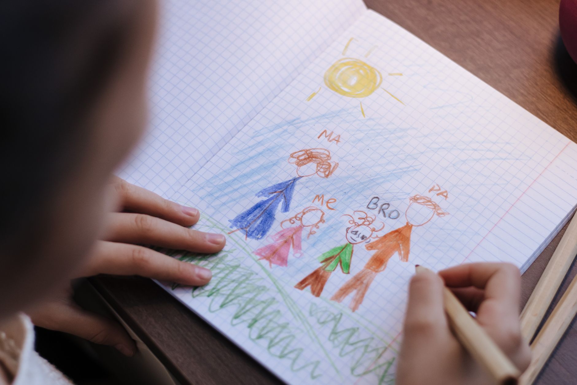 A child drawing a family portrait with crayons in a notebook on a wooden table.