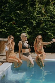 Three women in bikinis relaxing by a pool, taking a selfie with a smartphone.