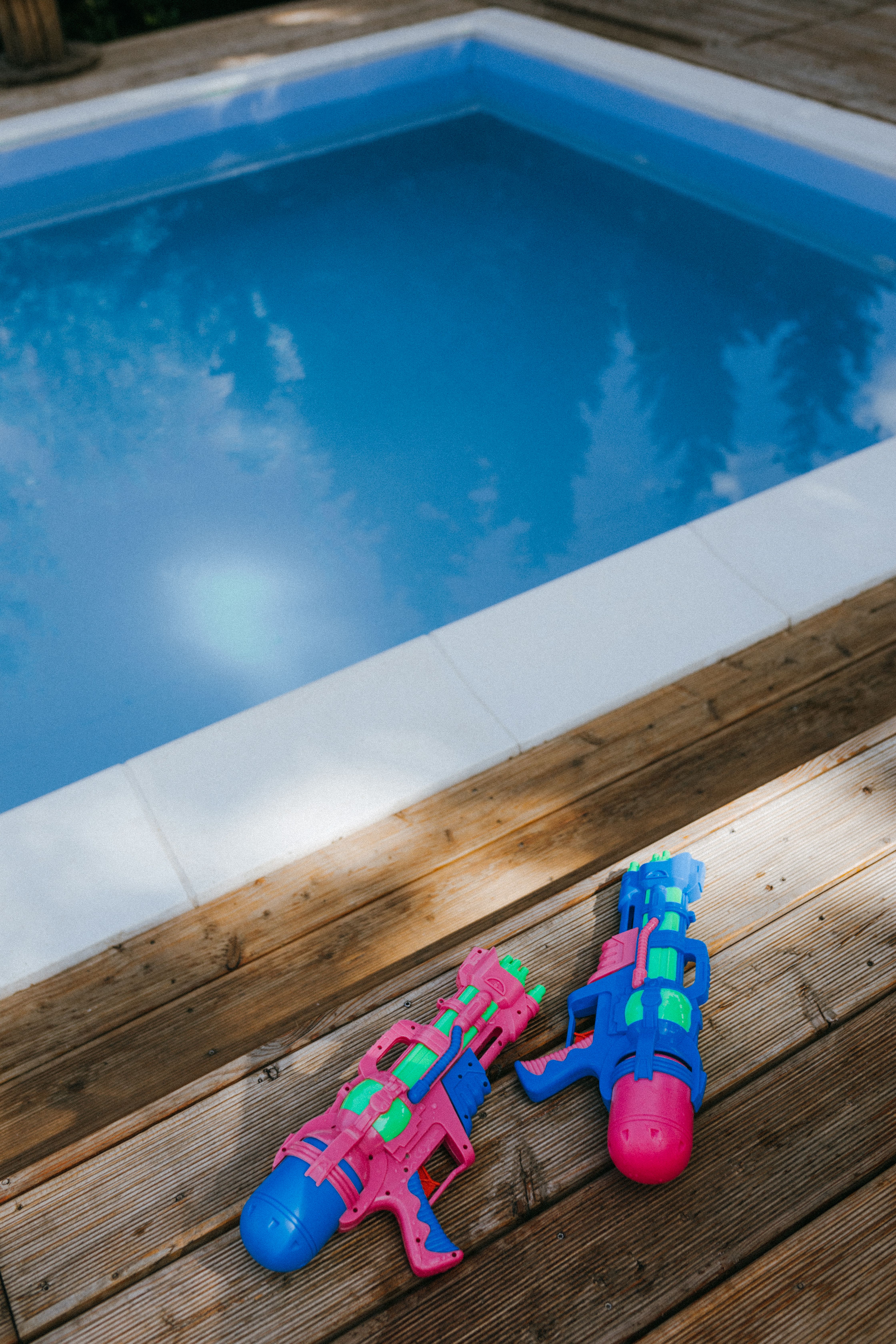 Colorful toy water guns on a wooden deck by a serene pool, perfect for summer fun.