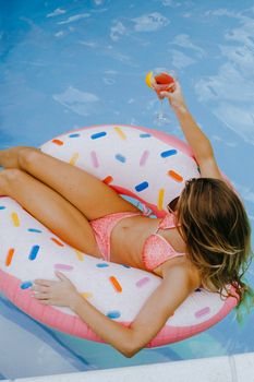 A woman in swimwear lounges on a donut float in a pool, holding a cocktail, enjoying summer leisure.