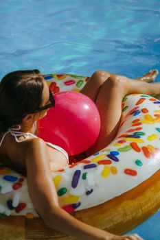 A woman sunbathing on a doughnut float in a pool, holding a pink ball and enjoying summer.