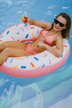 Smiling woman in bikini enjoying a cocktail on a floating donut tube in a swimming pool.