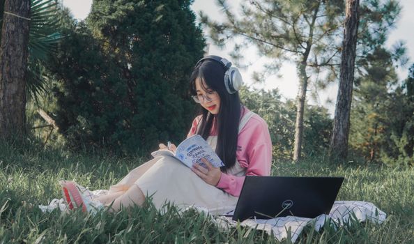 Asian woman studying outdoors with headphones, notebook, and laptop on the grass under trees.