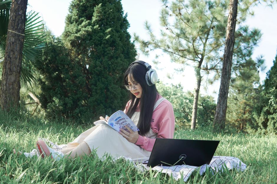 Asian woman enjoying outdoor study session with laptop and headphones, surrounded by nature.