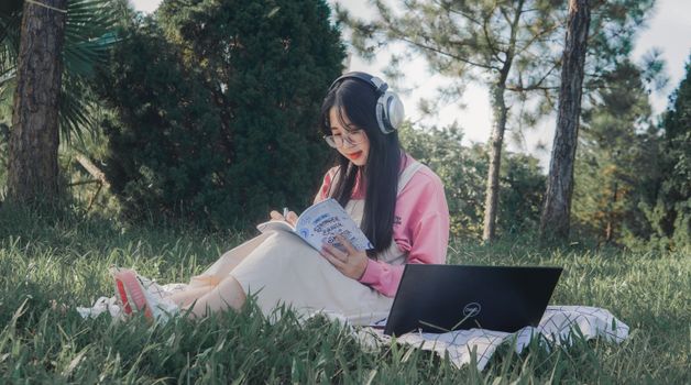 Asian woman studying in a park with headphones and laptop, enjoying a sunny day.