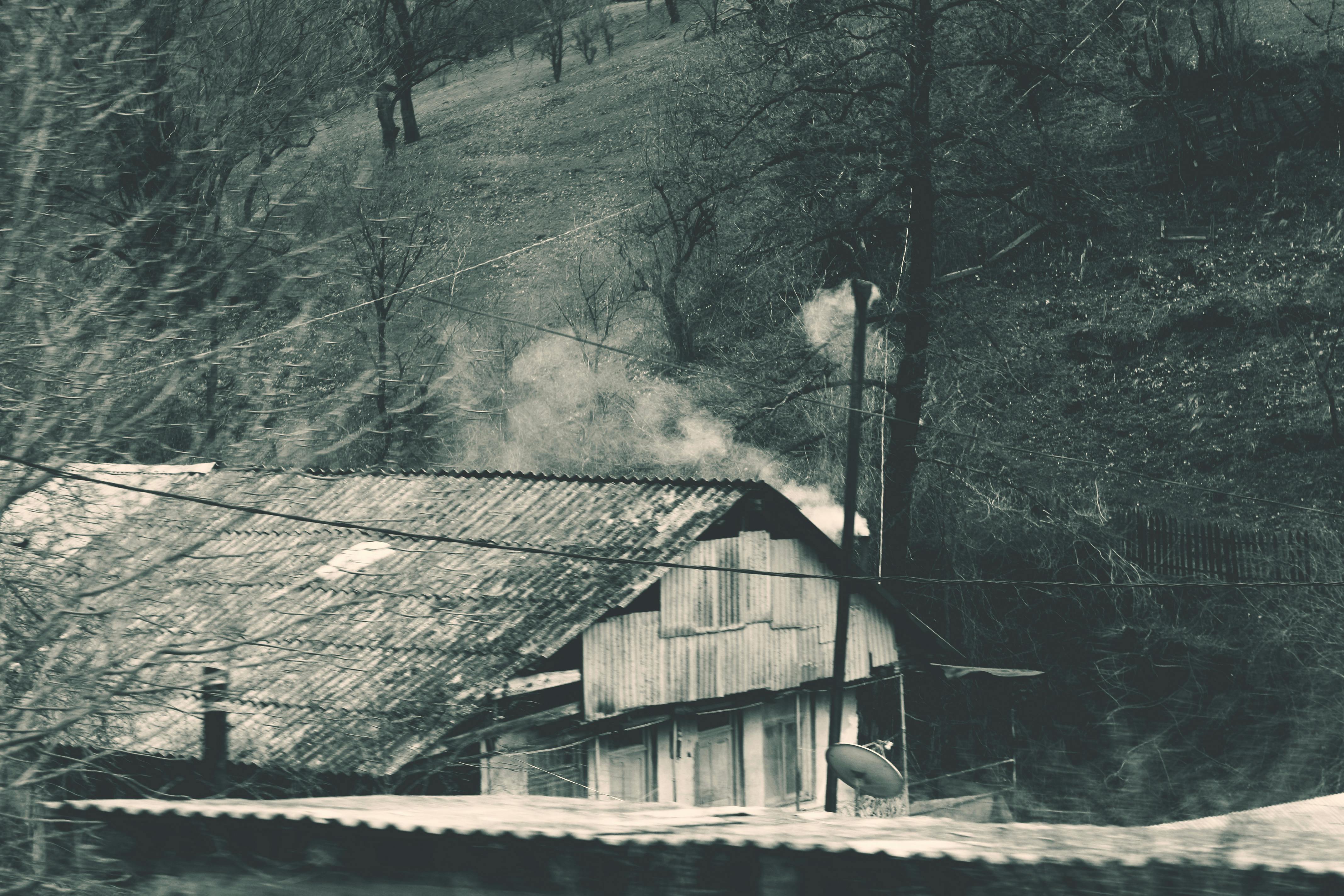 一幅黑白图像中，烟雾从乡村小屋升起，背景是迷雾笼罩的森林