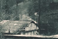 一幅黑白图像中，烟雾从乡村小屋升起，背景是迷雾笼罩的森林