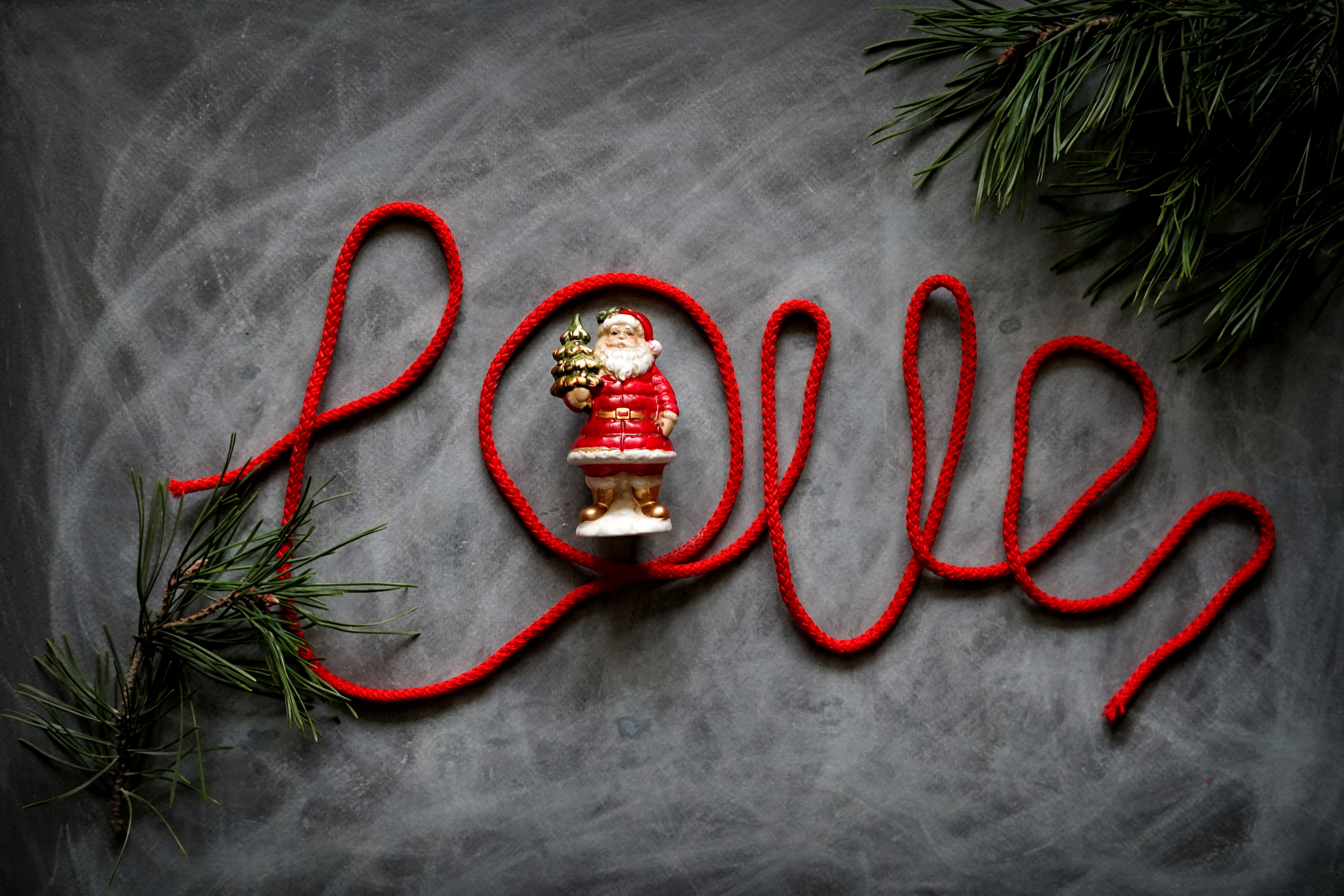 Flat lay of a Santa Claus figurine surrounded by pine branches and red rope spelling 'Love.'