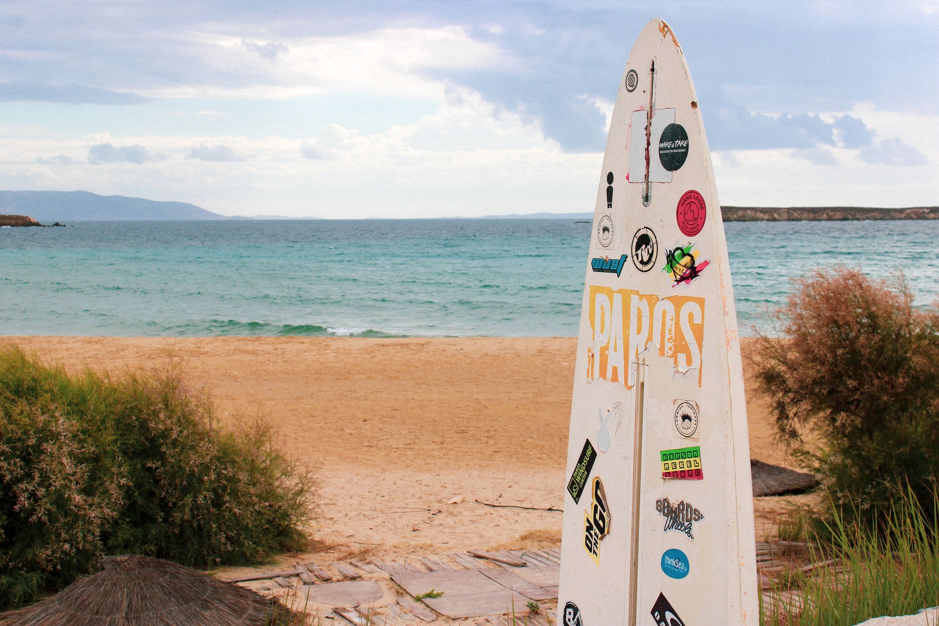 A surfboard with stickers on Paros beach with cloudy skies and a tranquil sea.