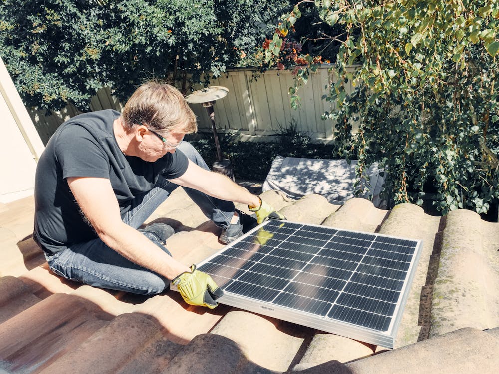 Free A Man in Black Shirt Sitting on the Roof while Holding a Solar Panel Stock Photo