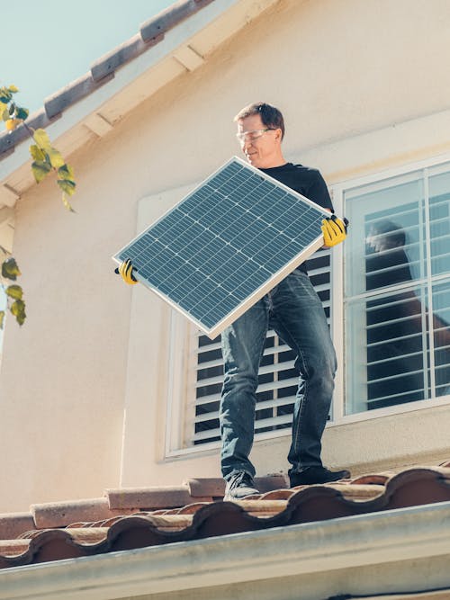 Free A Man in Black Shirt Standing on the Roof while Holding a Solar Panel Stock Photo