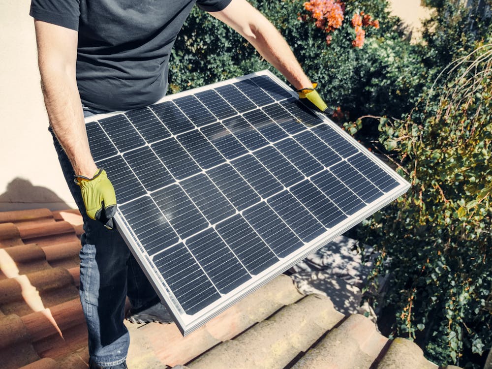 Free A Person in Black Shirt Holding a Solar Panel while Standing on the Roof Stock Photo