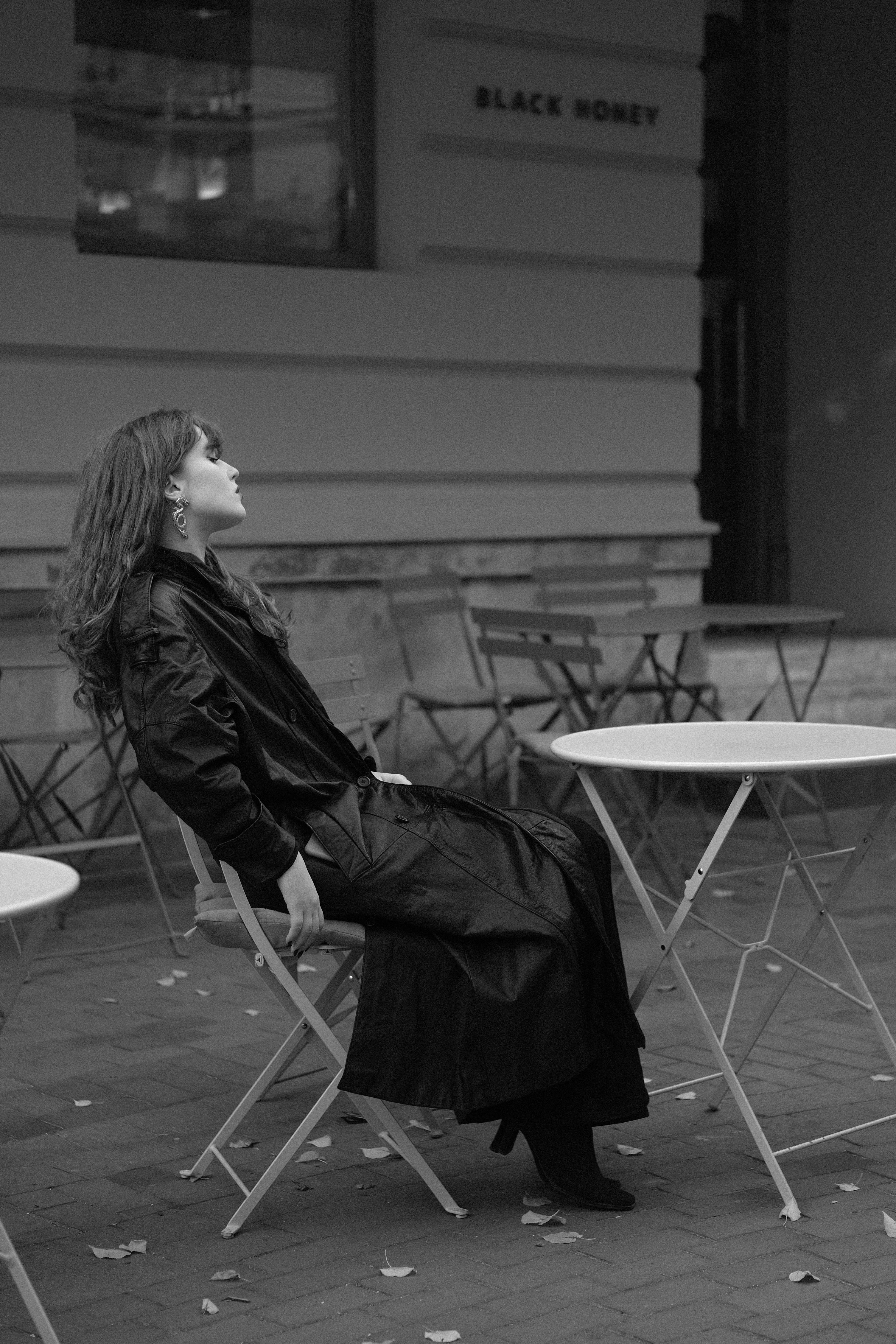 A woman in a long jacket sitting outdoors at a cafe, captured in black and white.