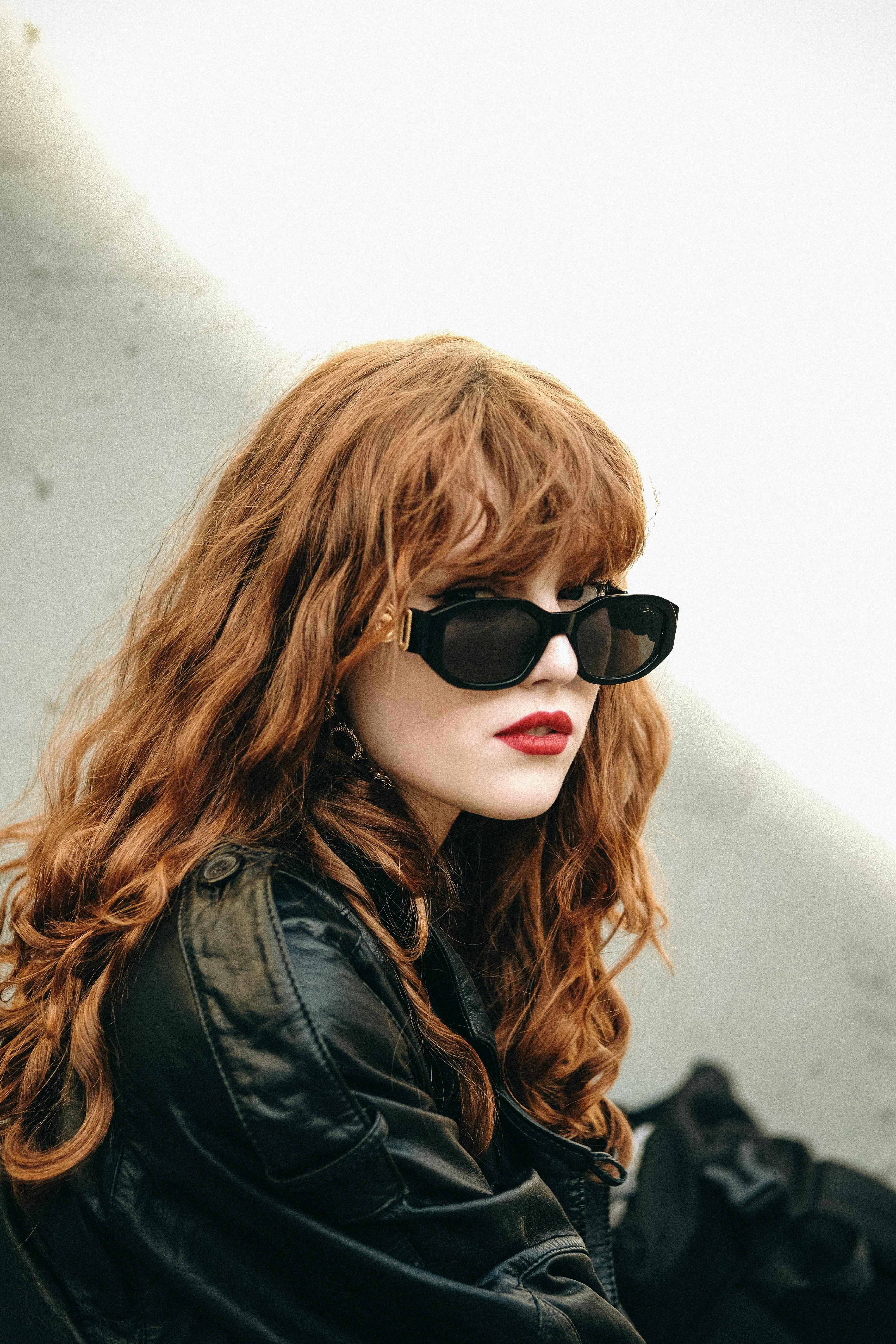 Fashionable woman with red hair in a leather jacket and sunglasses, looking stylish outdoors.