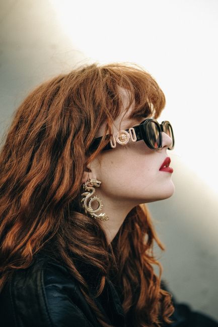 Stylish red-haired woman posing in sunglasses and fashionable earrings against a muted background.