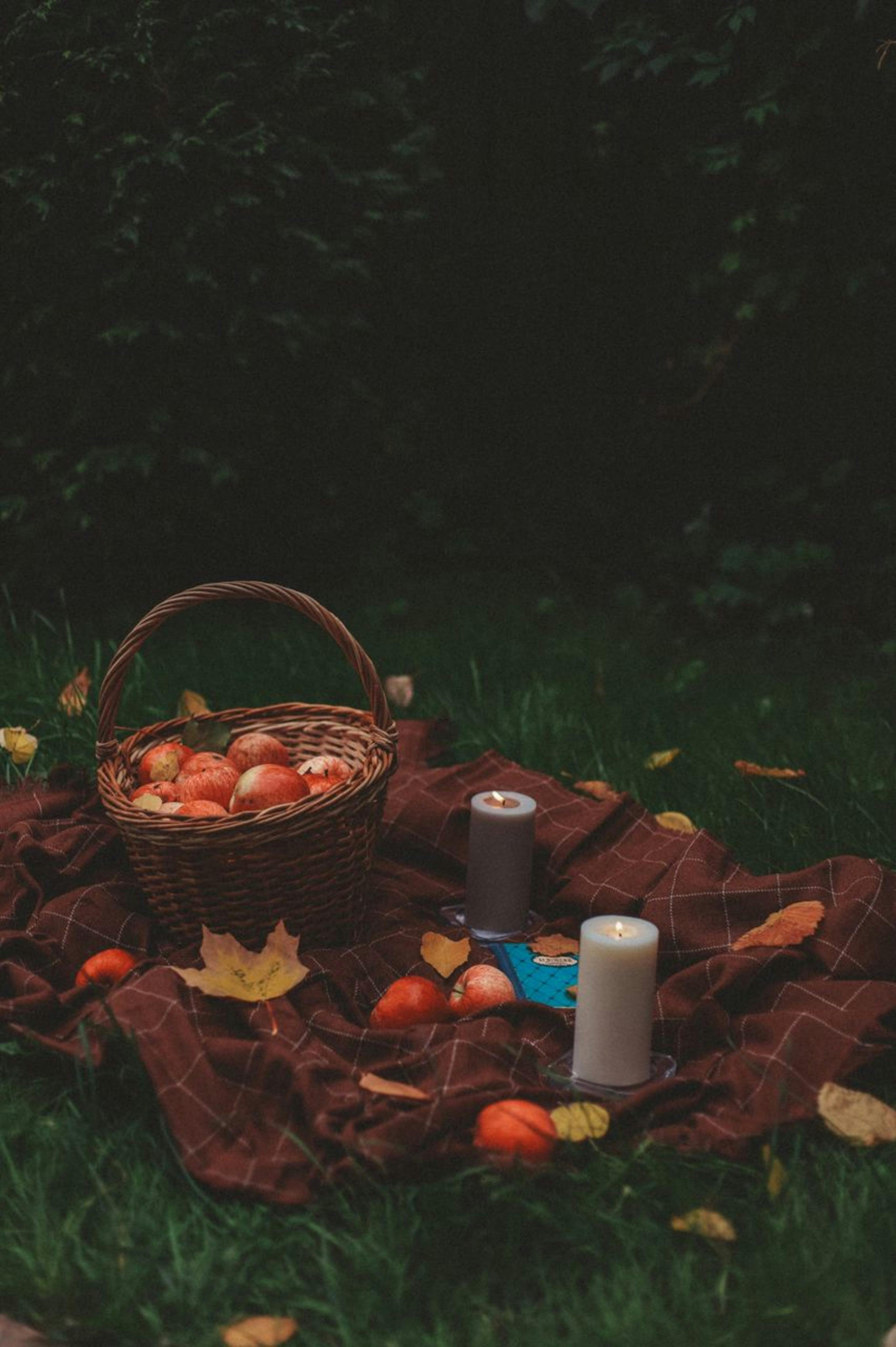 Autumn picnic scene with apples, basket, and candles on a blanket outdoors.