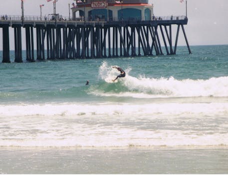 A surfer catches a wave near the iconic Huntington Beach Pier on a sunny day.