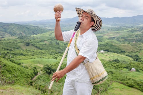 Man Playing a Flute, Holding a Rattle and Standing on the Background of a Green Valley 