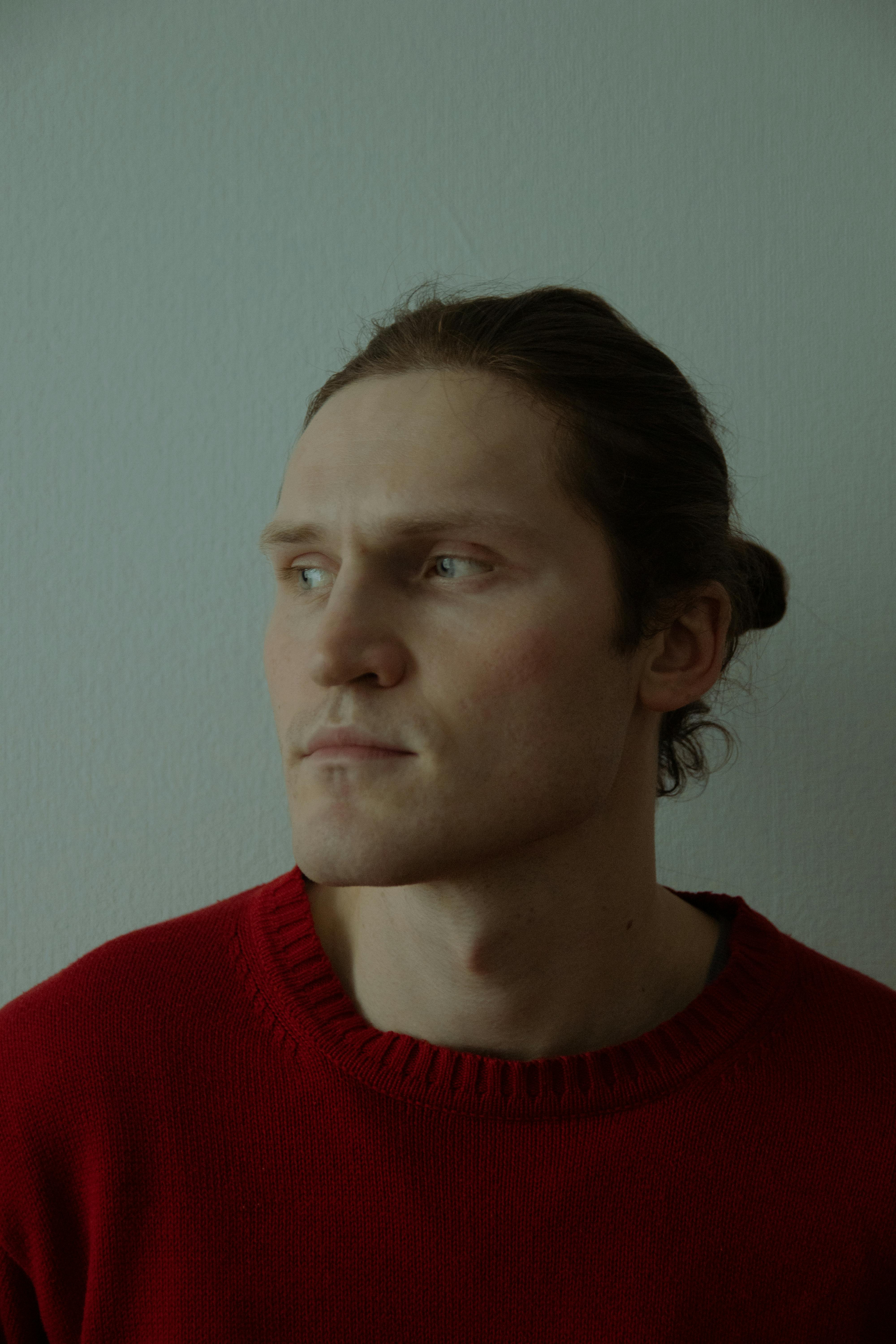 A serene portrait of a man with a hair bun wearing a red sweater, captured in a contemplative mood indoors.