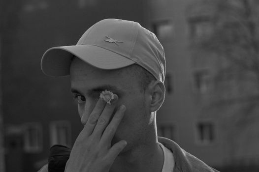 Monochrome photo of a man wearing a cap, holding a flower near his eye, with a selective focus effect.