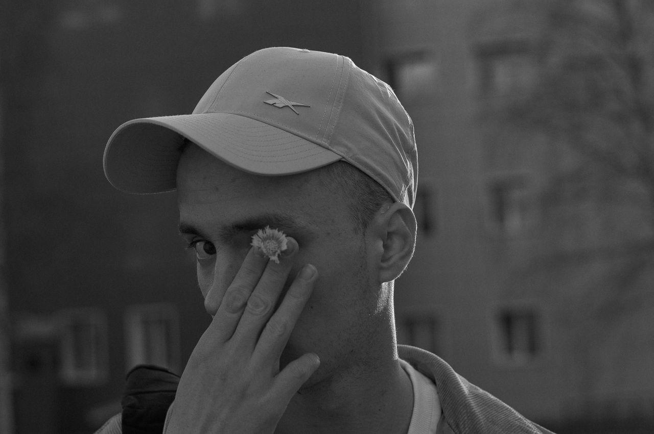 monochrome photo of a man wearing a cap holding a flower near his eye