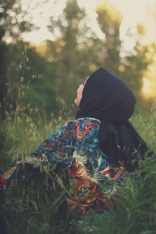 Free Woman wearing a hijab in a floral dress sitting peacefully in a grassy outdoor setting. Stock Photo