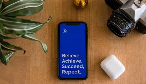 Free Flat lay of smartphone displaying motivational text alongside camera and AirPods on a wooden surface. Stock Photo