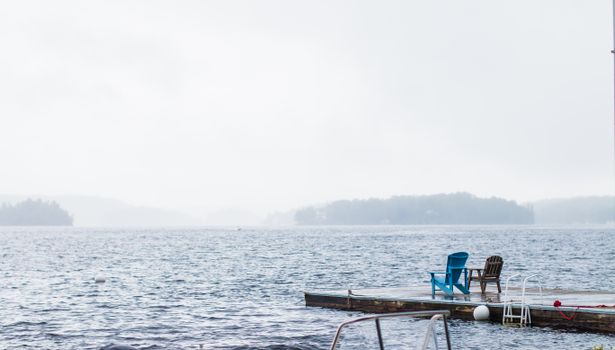 Empty wooden chairs left on pier with amazing view on calm lake on misty day