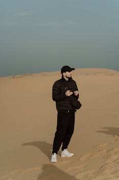 A man stands on sandy desert dunes, holding photography equipment, wearing black clothing.