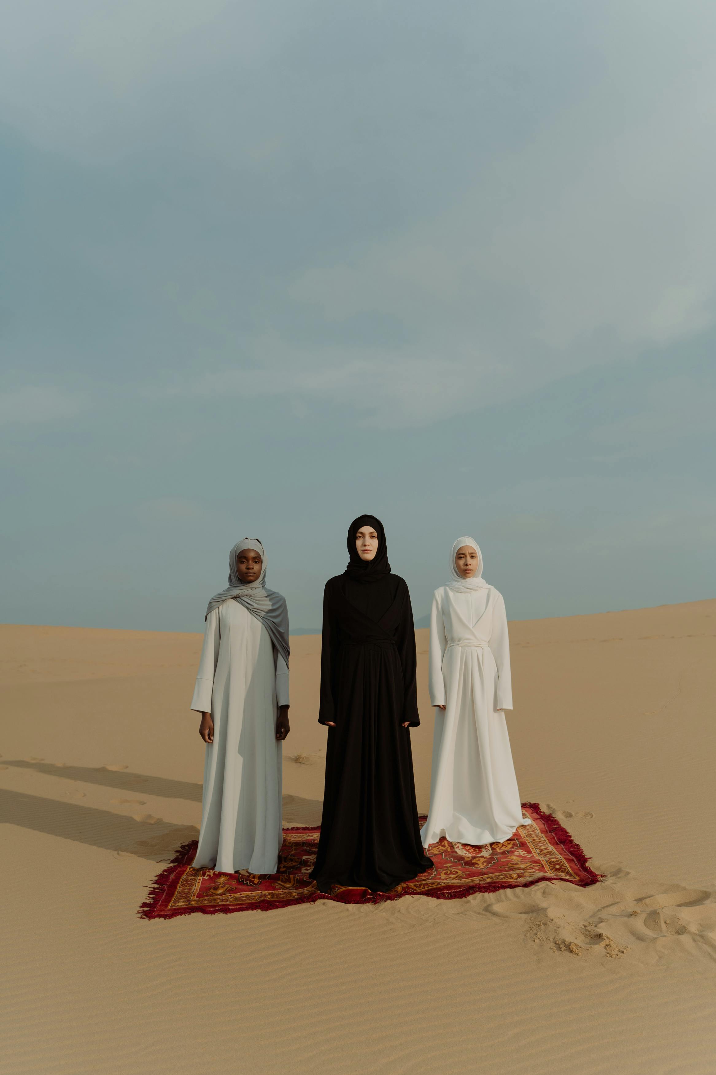 Three women wearing hijabs standing on a red carpet in a vast desert under a blue sky.
