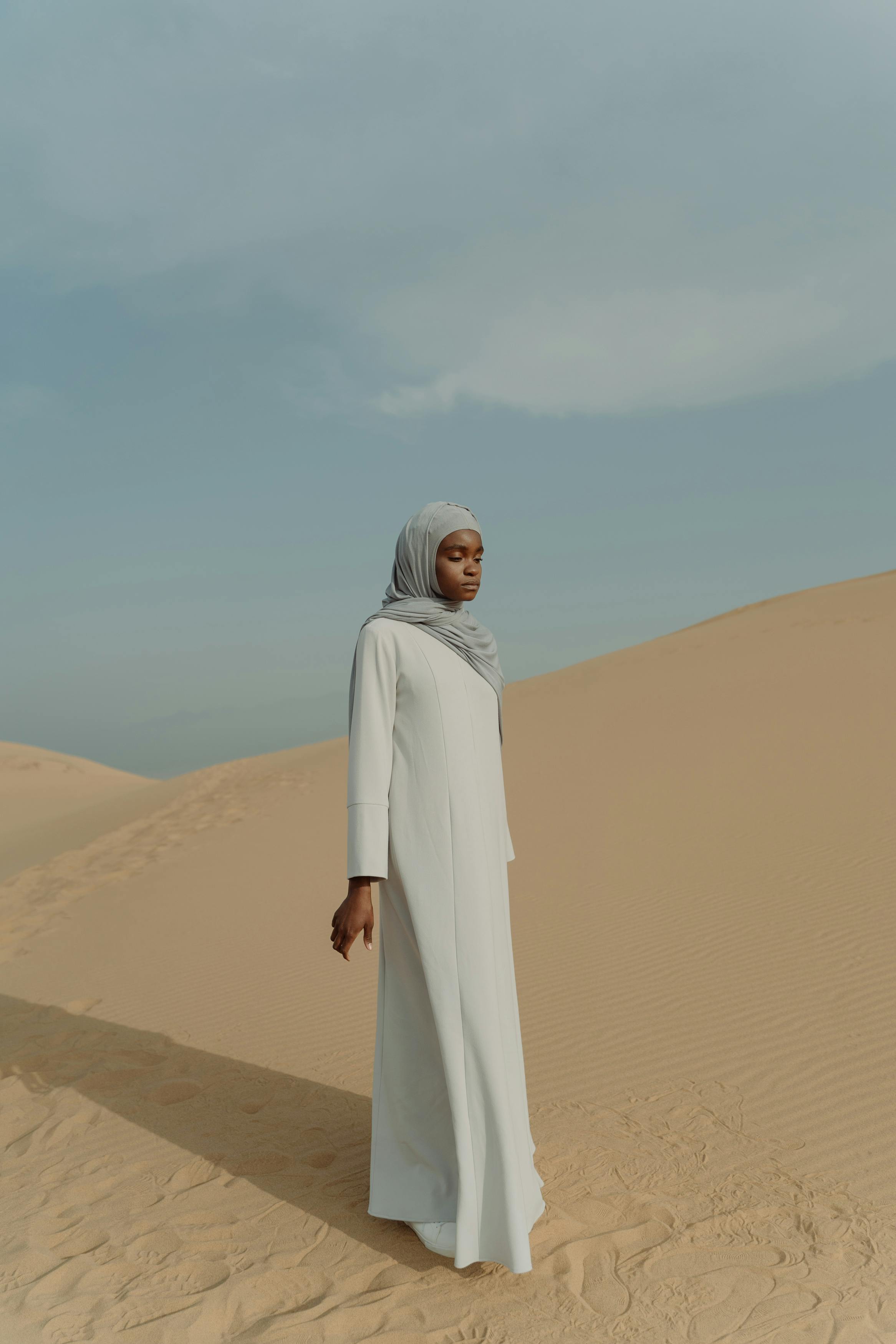 A woman in a hijab gracefully walks through vast desert sand dunes under a clear sky.