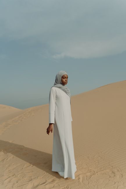 A woman in a hijab gracefully walks through vast desert sand dunes under a clear sky.