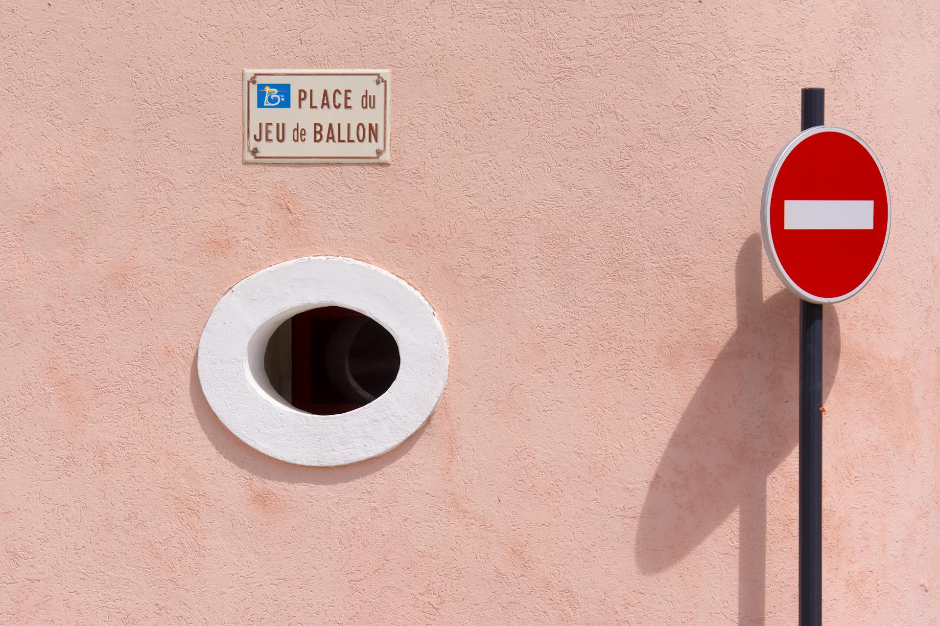 Simple urban scene featuring a no entry sign beside a circular window and a street name plaque.