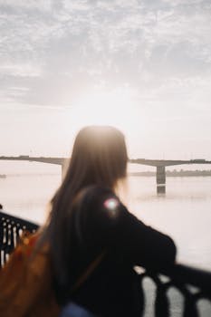 Silhouette of a woman with a backpack gazing at a peaceful river and bridge during sunset.