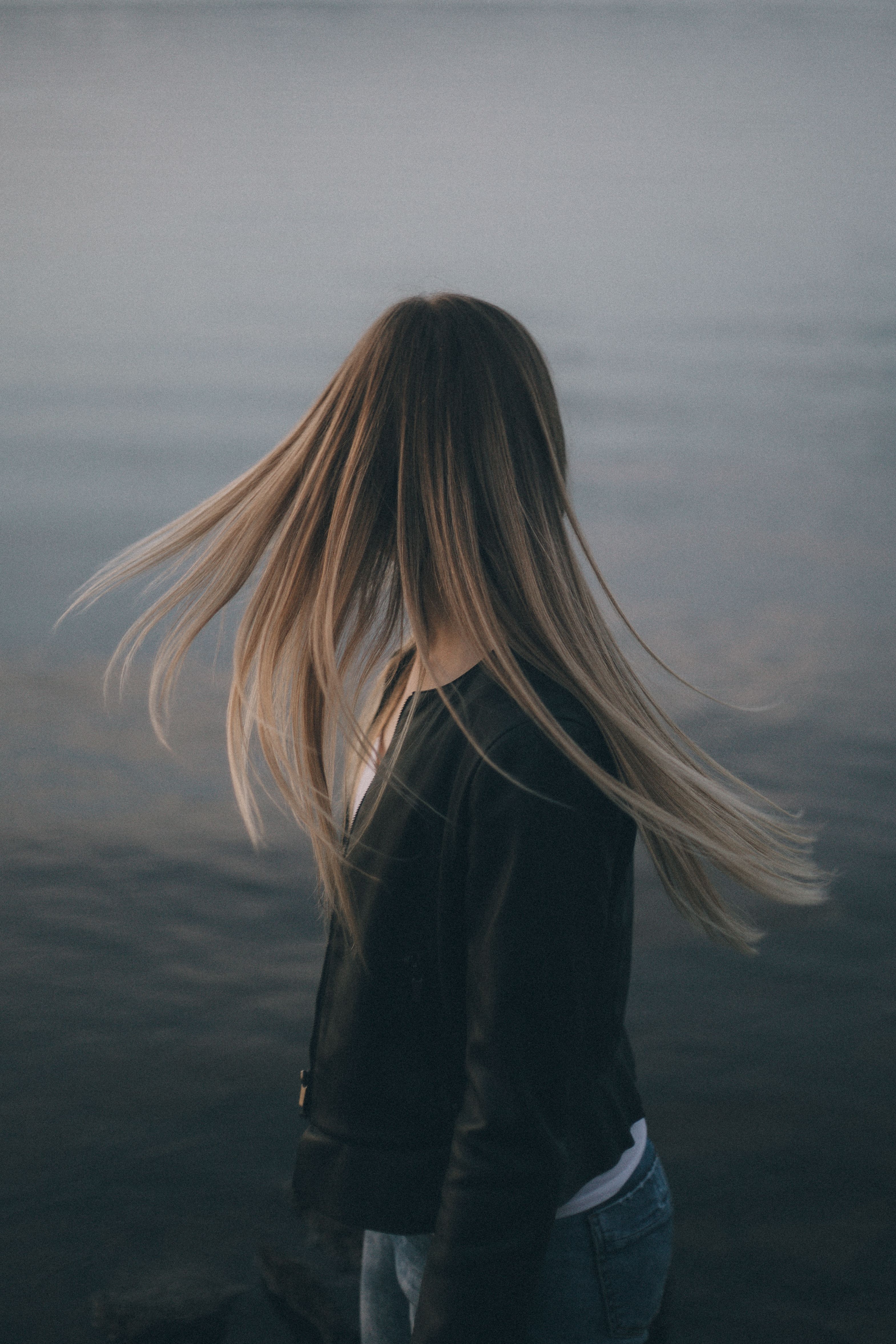 A woman with long blonde hair stands by the river at sunset, gazing away.