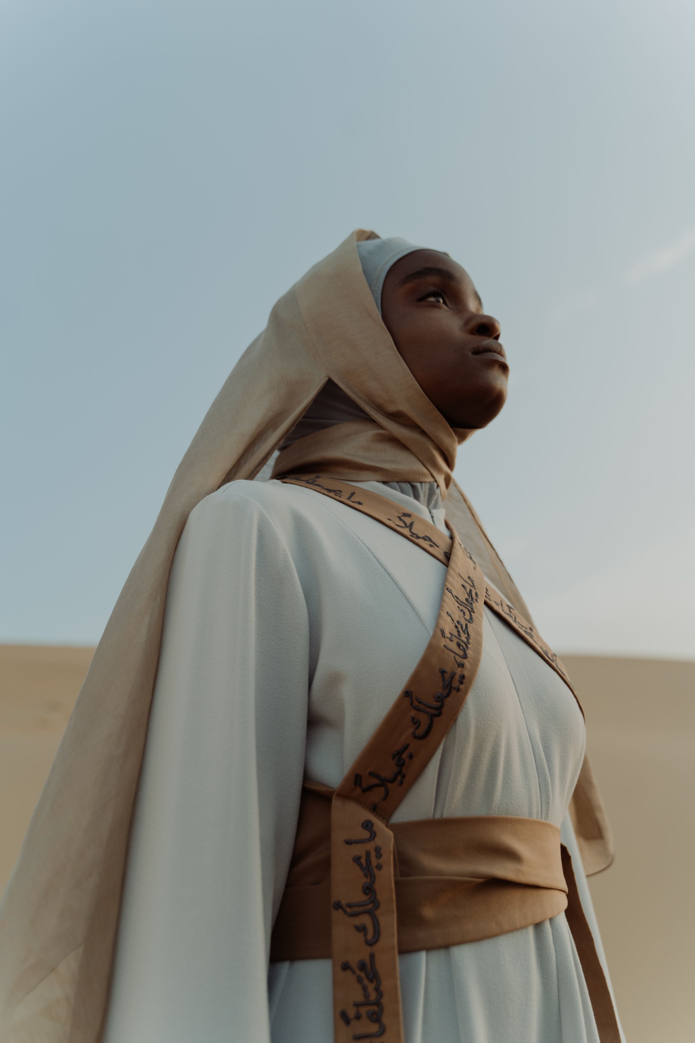 A woman in a stylish hijab and robe poses gracefully against a serene desert background.