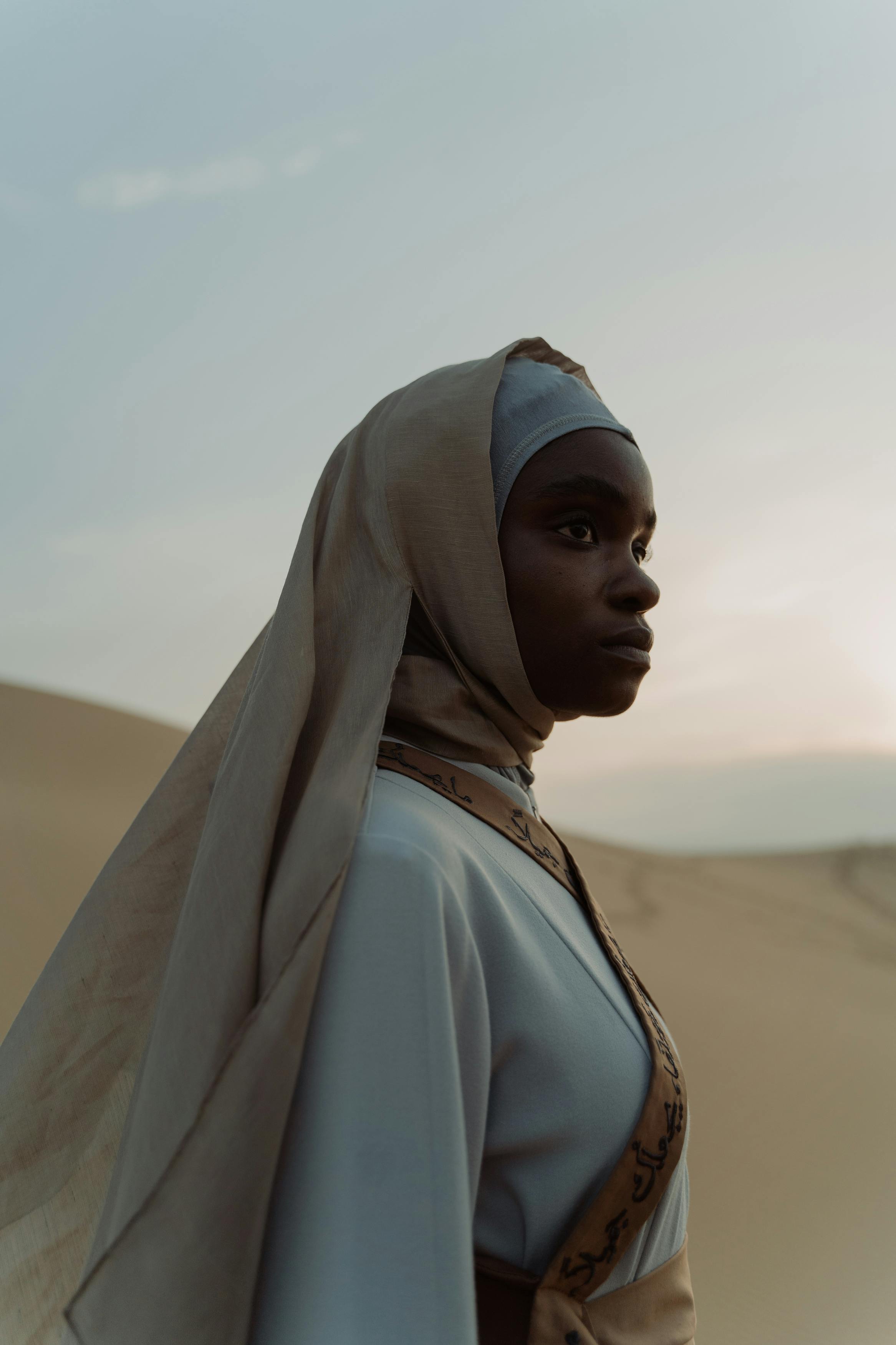 Side view of a woman in a hijab standing in a serene desert environment at dusk.