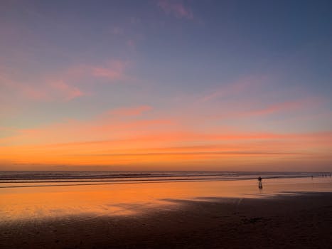 A tranquil beach scene at sunset with vibrant orange and pink hues reflected on the water.