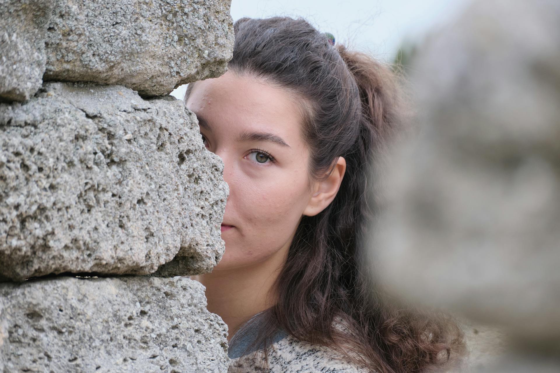 A woman with long hair peers past a stone wall, partially hiding her face, in an outdoor setting.