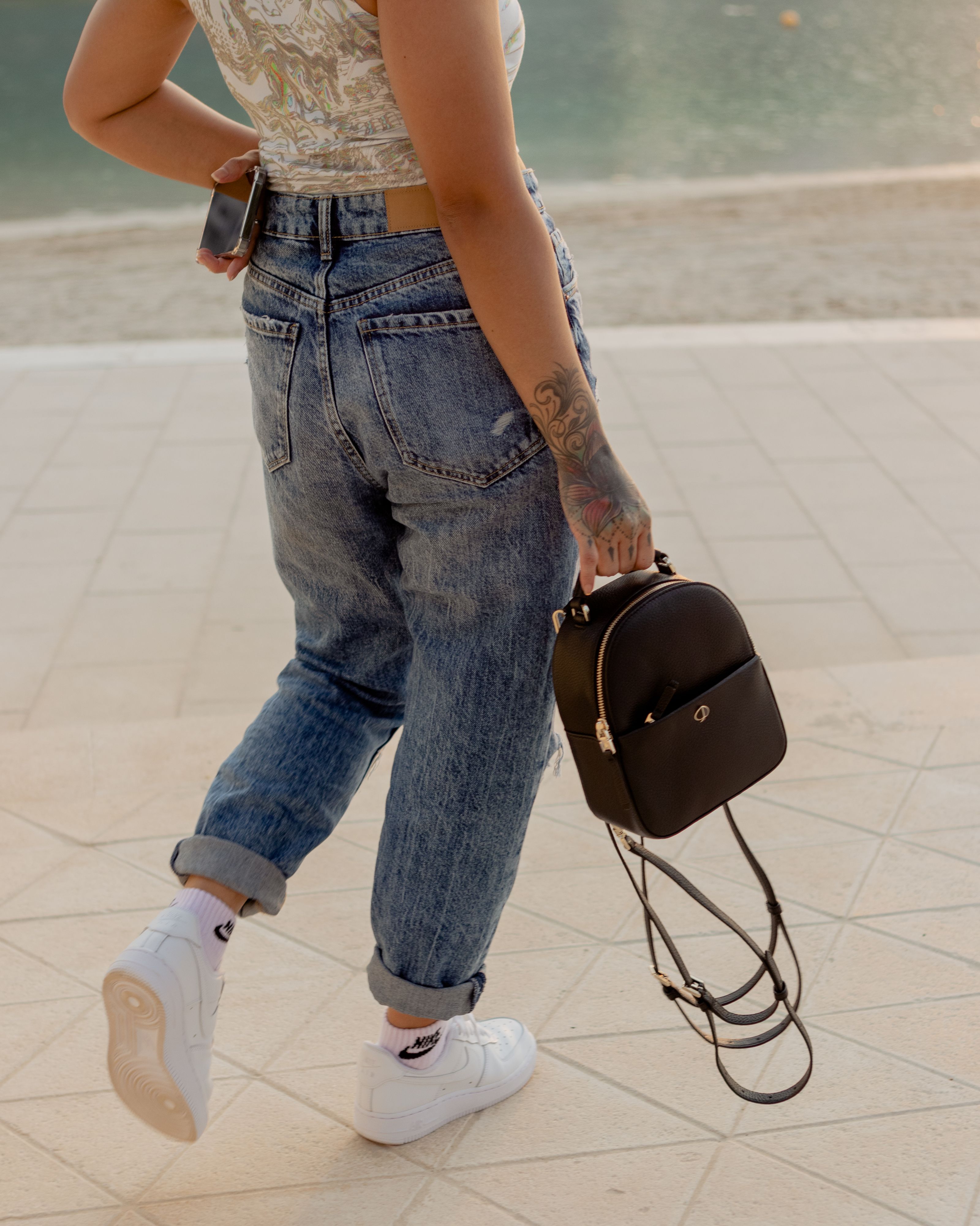 Uma jovem mulher fashion com tatuagens caminha ao ar livre segurando uma mochila preta. Estilo casual e moderno.