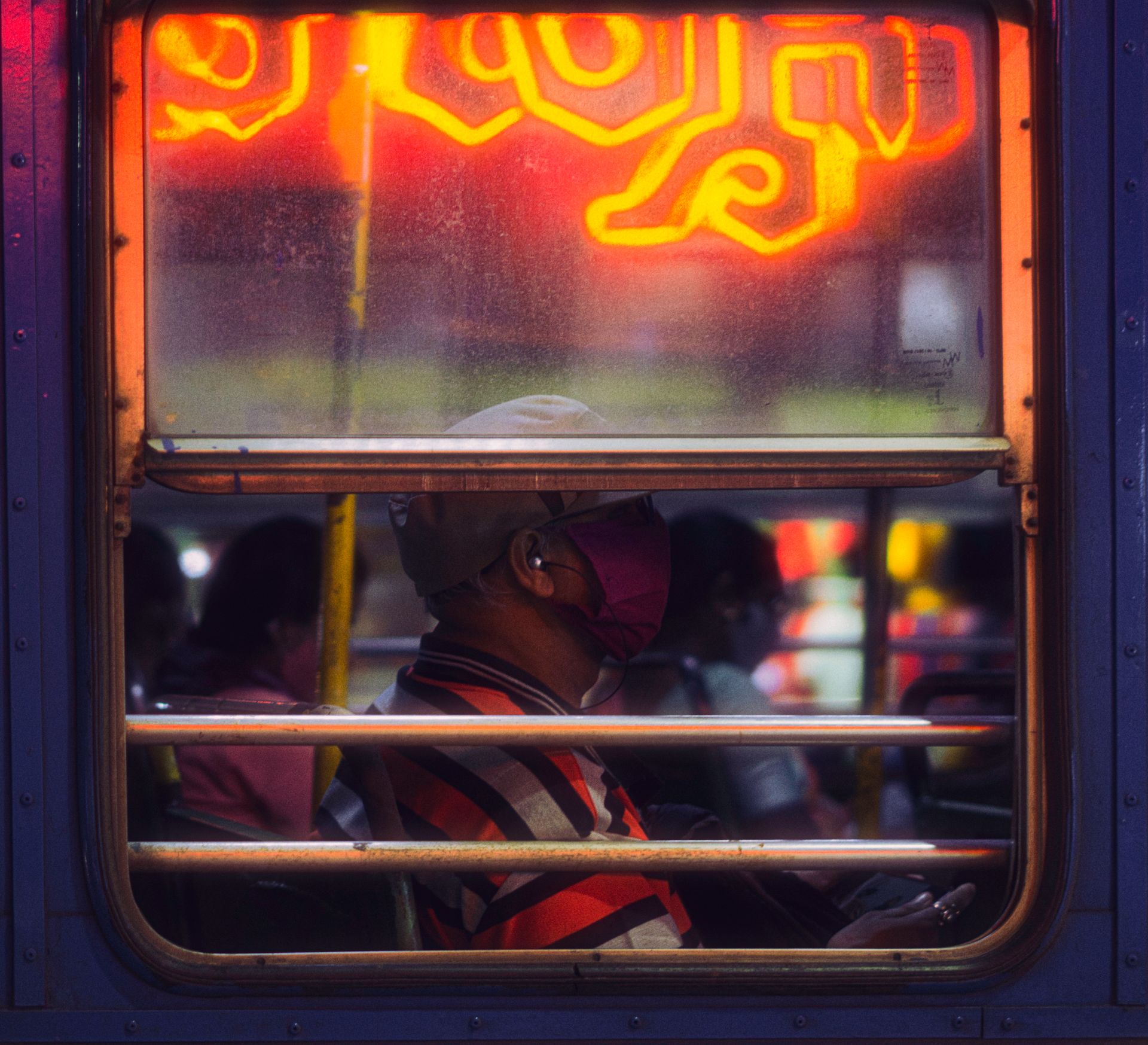 A man in a mask sits on a bus in Pune, India, under vibrant neon lights.