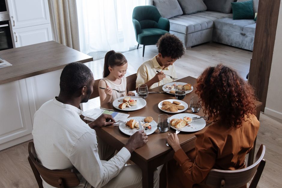 Dark-skinned Nigerian family of four — mother in adire blouse, father in plain kaftan, two young children — sitting around a modest wooden table in a simple Lagos home kitchen, sharing a small pot of vegetable soup and eba, expressions thoughtful and tired, natural window light casting soft shadows, photorealistic