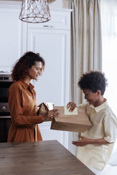 A mother hands her son a lunch bag in the kitchen, both smiling warmly.