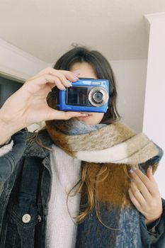 A woman holds a blue camera indoors, partially covered with a colorful scarf, capturing a unique self-portrait.