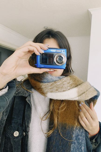 A woman holds a blue camera indoors, partially covered with a colorful scarf, capturing a unique self-portrait.