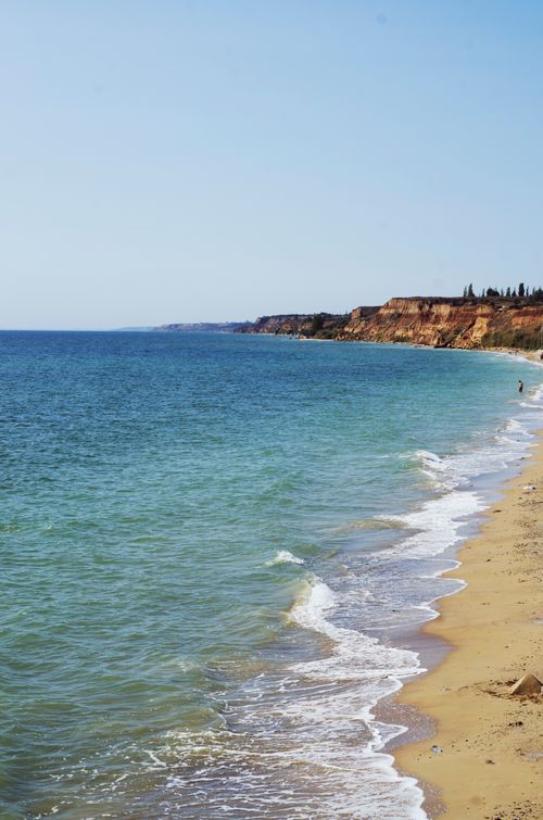 Free Beautiful coastal landscape featuring a sandy beach, ocean waves, and distant cliffs under a clear blue sky. Stock Photo
